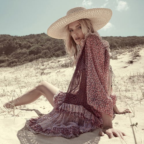 A woman sitting in the sand in a sheer coverup and a sunhat