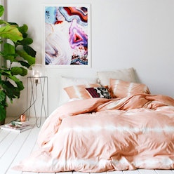 A bedroom with a bed with peach-white sheets, a painting, a plant, a nightstand and a lamp