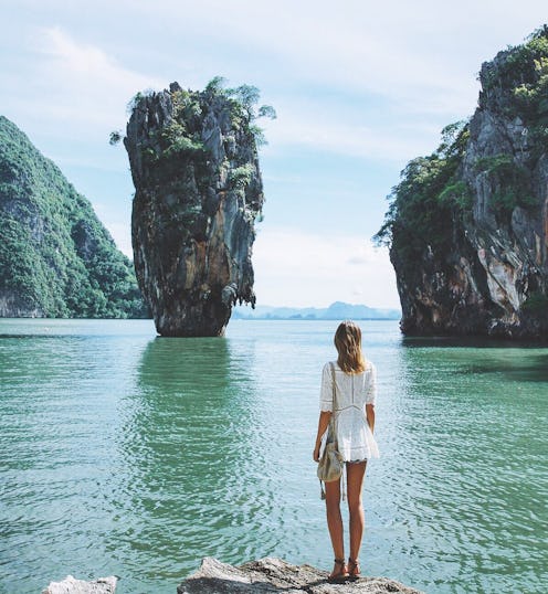 a woman on a solo vacation stands in front of a surface of water with rock formations sticking