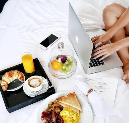 A woman sitting on a bed and using her laptop with breakfast plates surrounding her 