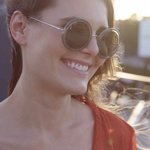 A woman with glowing skin and round sunglasses, smiling