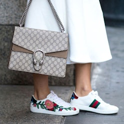 A closeup of a woman's feet in Gucci sneakers with a rose on the side and a Gucci bag