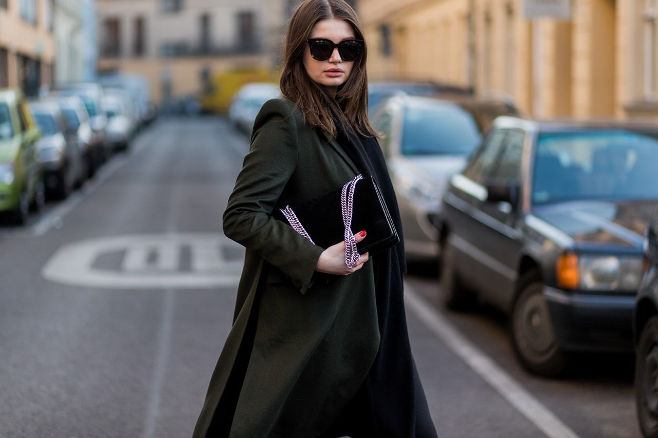 A woman in a long black coat standing in the middle of the street holding her black handbag