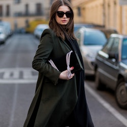 A woman in a long black coat standing in the middle of the street holding her black handbag