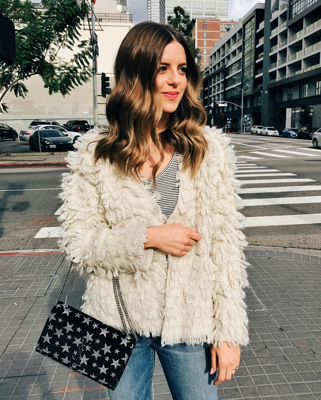 A woman with brown wavy hair standing outside in a puffy jacket and a purse with a star pattern