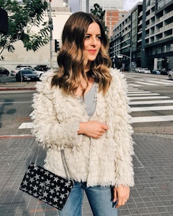 A woman with brown wavy hair standing outside in a puffy jacket and a purse with a star pattern