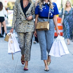 Two women walking down the street, one is wearing a glittery silver suit, the other is in a blue top...