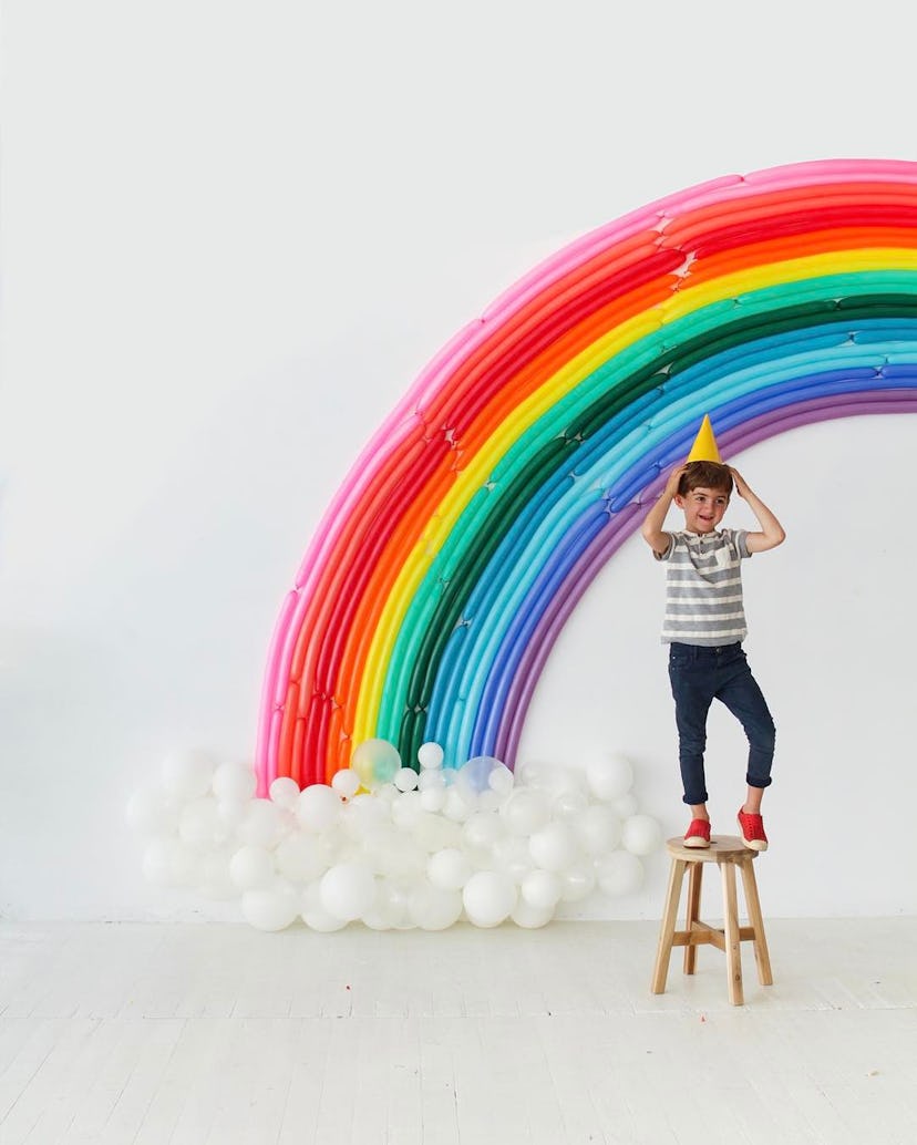 A boy wearing a party hat standing on a stool with a balloon rainbow behind him