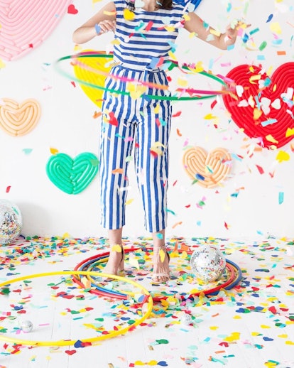 A girl wearing a striped jumpsuit spinning a hula-hoop with confetti falling on a floor around her