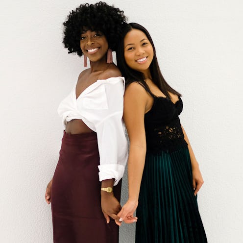 Two women standing back to back holding hands and smiling, one is in a white button up shirt and the...