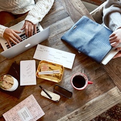 Two people at a wooden table working on their laptops, with coffee and notebooks at the center 