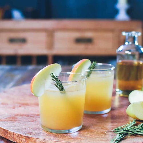 Two glasses with the Rosemary Mezcal Apple Cider, apple slices, rosemary leaves and two bottles