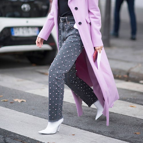 A woman crossing a street with denim pear-beaded jeans, a lavender coat and white boots