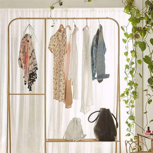 A gold clothing rack in front of white curtains and a wall covered in plants is full of tops and han...