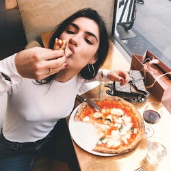 A brunette woman in a white shirt eating pizza with her hands and looking up