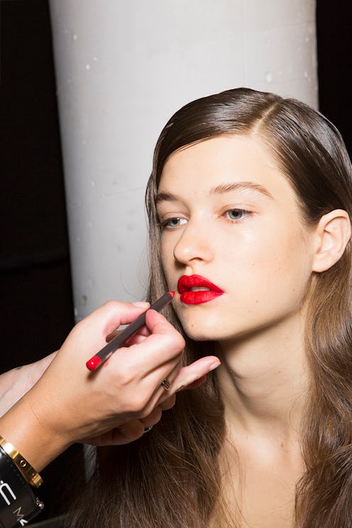 A model sitting and getting red lipstick applied to her by a makeup artist