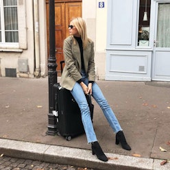 A blonde woman sitting on a suitcase on the street, wearing it-girl jeans, a black turtleneck, beige...