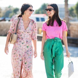 A girl in a maxi dress walking next to her friend in green pants and a pink t-shirt, both wearing Tr...