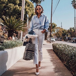 A woman in a blue button-up shirt and white pants walking down the sidewalk during the summer