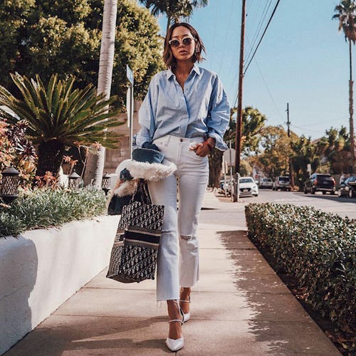 A woman in a blue button-up shirt and white pants walking down the sidewalk during the summer