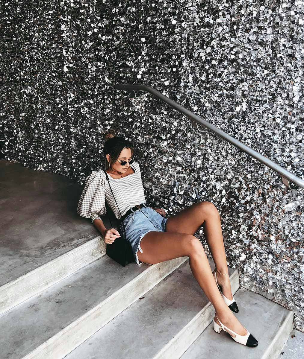 A woman in a big blouse in white, blue denim shorts, black-white ballet flats, and a black bag, sitt...