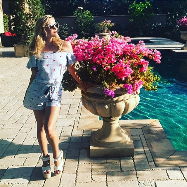 Reese Witherspoon in her home in a blue romper, next to a large potted plant 