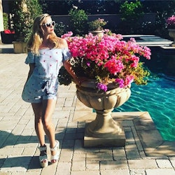 Reese Witherspoon in her home in a blue romper, next to a large potted plant 