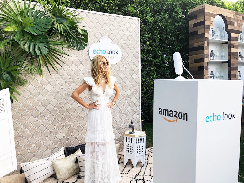 A blonde woman standing next to an Echo Look Amazon promo stand while wearing a white dress