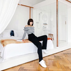 A woman wearing a grey top and black pants posing on a large bed thats next to a see-through shower