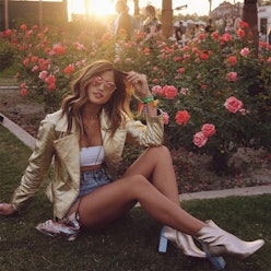 A woman sitting in front of a rose garden wearing a gold jacket, jean shorts and gold pumps