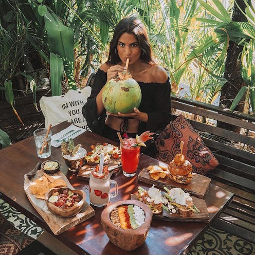 A woman sitting on a bench with a table in front of her full of superfoods while she drinks out of a...