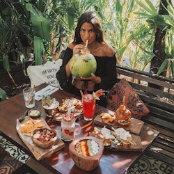 A woman sitting on a bench with a table in front of her full of superfoods while she drinks out of a...