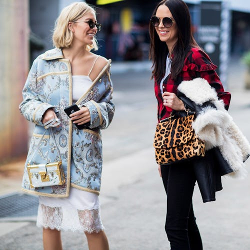 Two women on the street, one in a blue jacket and white bag and the other in a plaid jacket and leop...