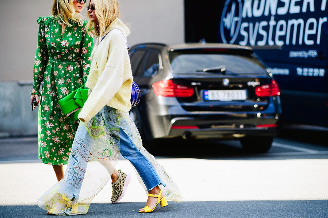 Two women walking at a parking lot, one in a hoodie with jeans and heels and the other in a green dr...