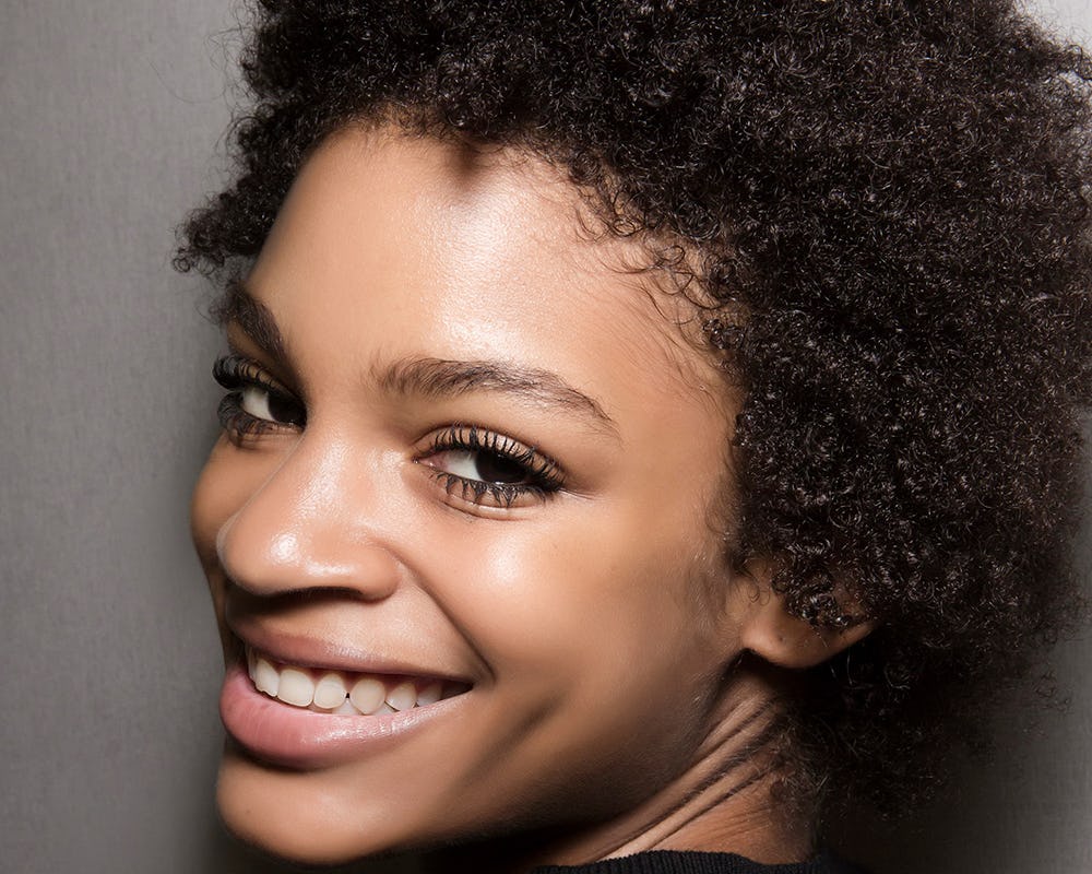 A model with curly brown hair smiling at the camera after applying a face oil