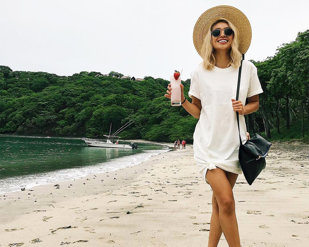 A woman in a white T-shirt-dress, large hat, sunglasses and a black bag holding a drink while standi...