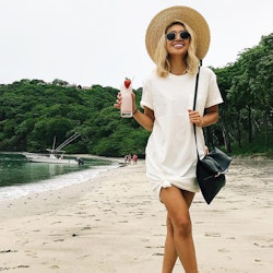 A woman in a white T-shirt-dress, large hat, sunglasses and a black bag holding a drink while standi...
