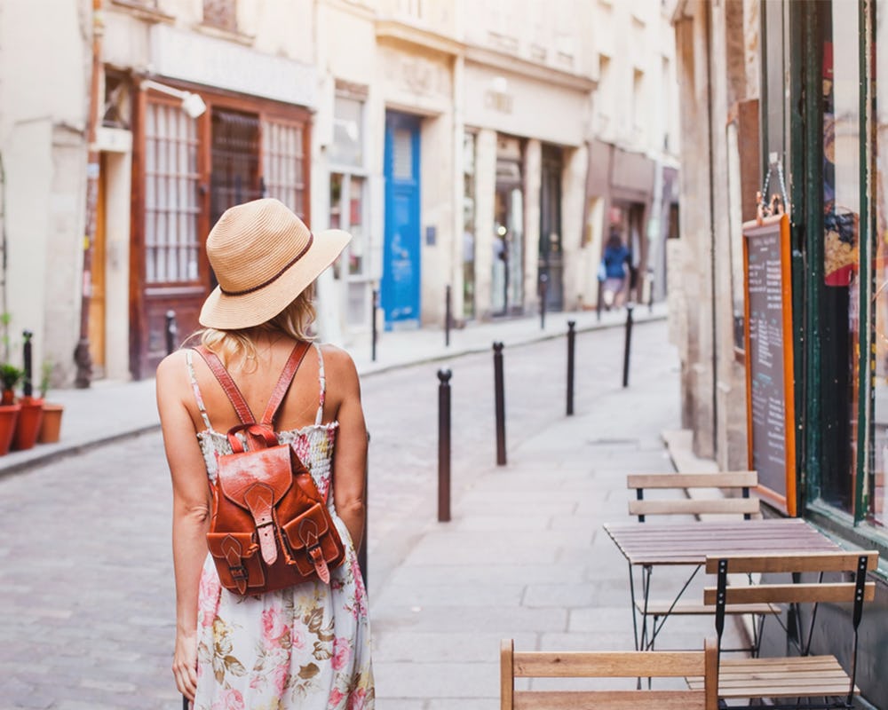 The back of a woman in a floral dress, straw hat and a small brown leather backpack on vacation