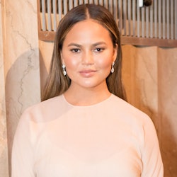Chrissy Teigen in a peach colored top posing in front of a brown background