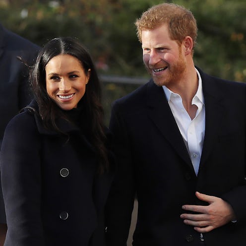 Meghan Markle in a black coat and Prince Harry in a black coat and a white shirt, smiling while hold...