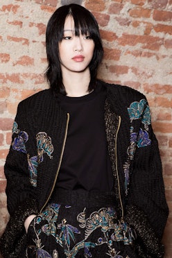 A black-haired model with a bob haircut in a black top, black floral jacket, and pants