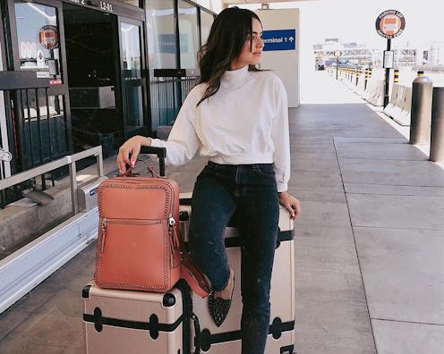A woman standing in front of the airport wearing a white turtleneck next to her budget-friendly lugg...