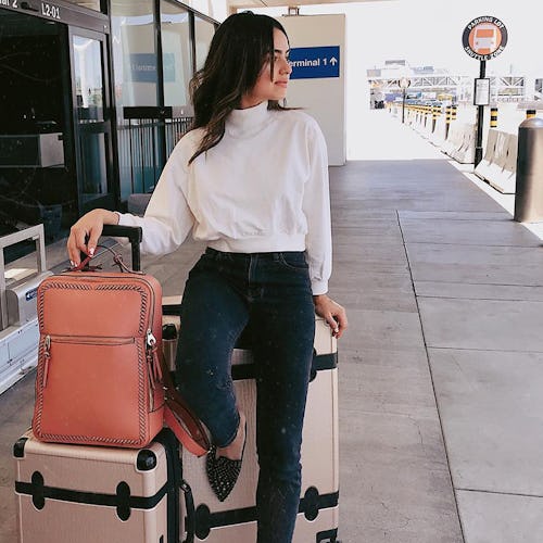 A woman standing in front of the airport wearing a white turtleneck next to her budget-friendly lugg...