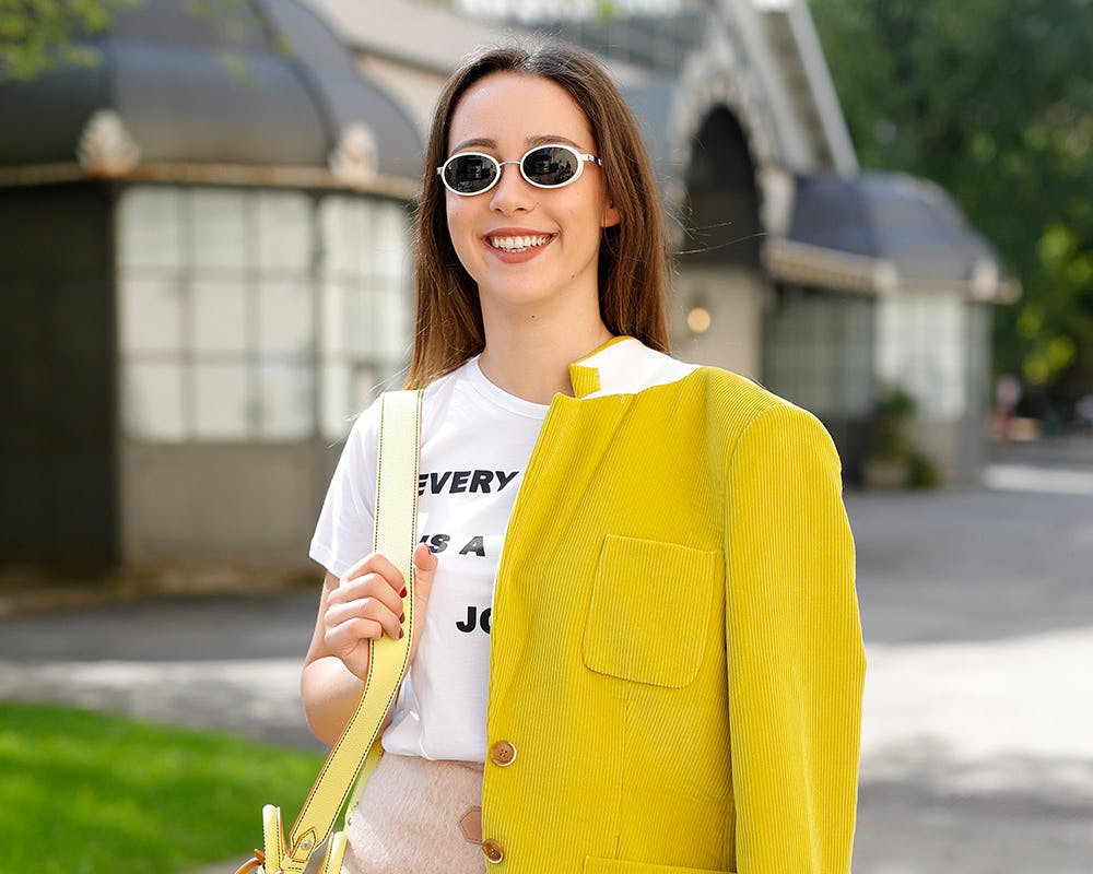 A woman in a white t-shirt, beige mini skirt and sunglasses with a yellow jacket draped over one sho...