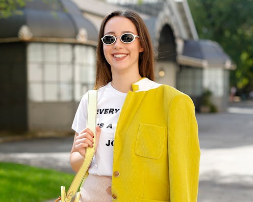 A woman in a white t-shirt, beige mini skirt and sunglasses with a yellow jacket draped over one sho...