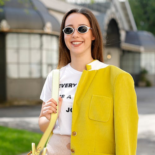 A woman in a white t-shirt, beige mini skirt and sunglasses with a yellow jacket draped over one sho...