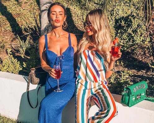 Two women sitting on a white wall, one is wearing a jean pantsuit and the other a striped pantsuit