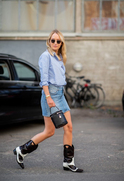 A woman walking in a blue shirt and blue denim skirt while carrying a black bucket bag