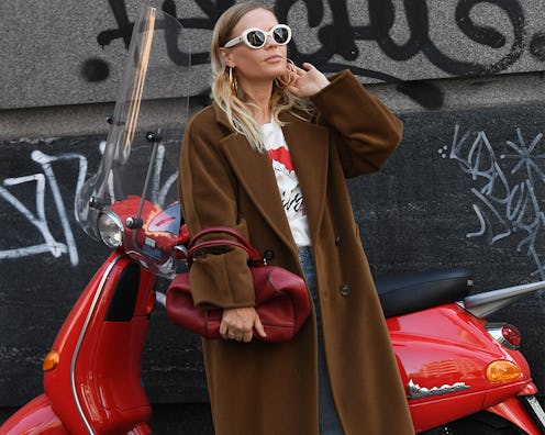 A woman posing in sunglasses, a brown coat, a white shirt and jeans in front of a red scooter