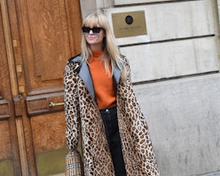 A blonde woman in an orange sweater, black pants, and a long leopard animal print coat 